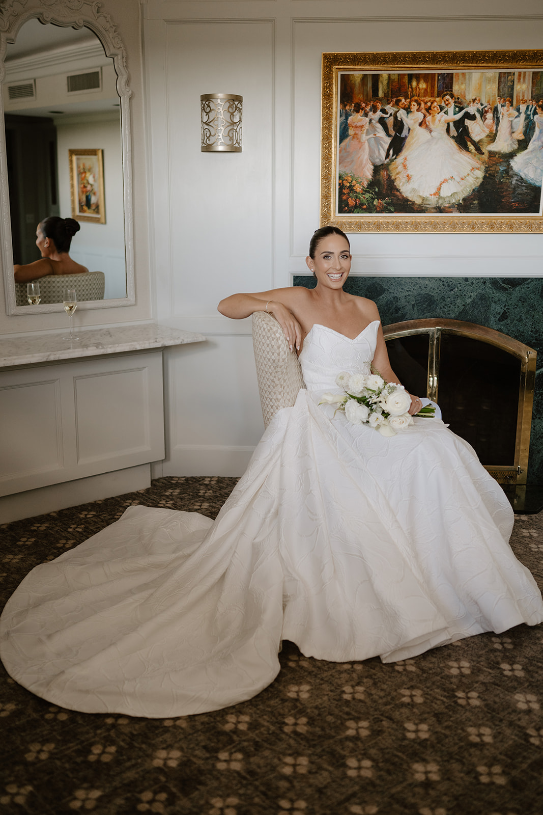 Smiling bride posing gracefully in her wedding gown, seated in a chair by a fireplace.